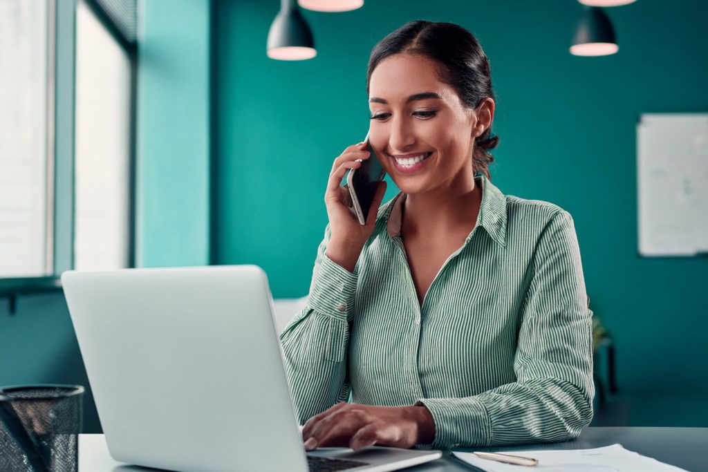 Photo of smiling confident woman, sitting at a laptop in her office, talking on a smartphone