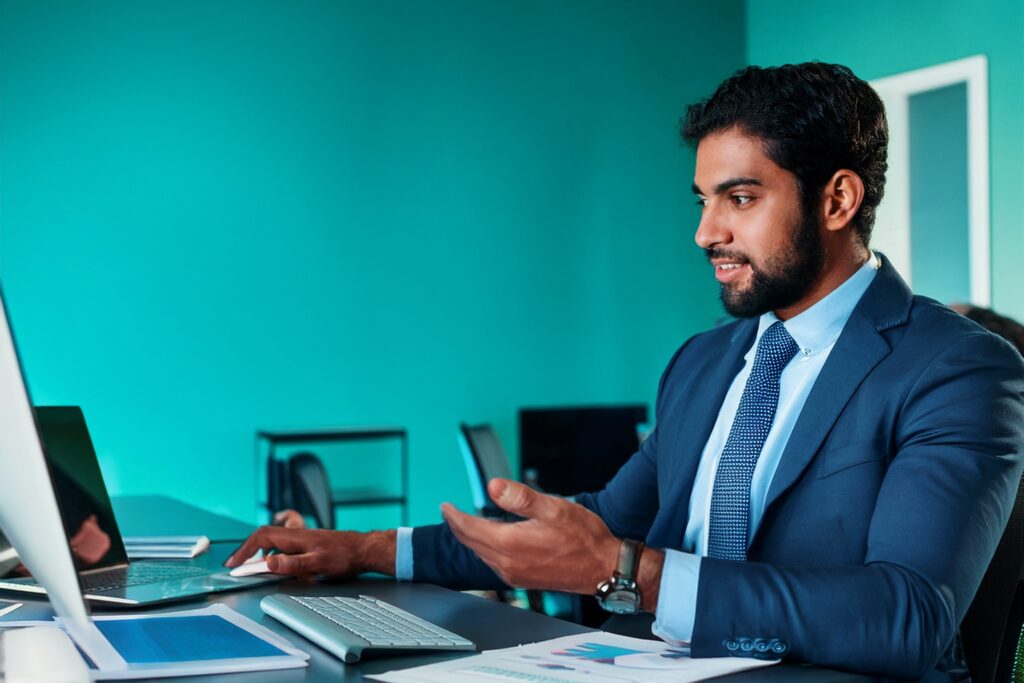 Business man in office with teal coloured walls showing colleagues somwthing on his desktop computer screen