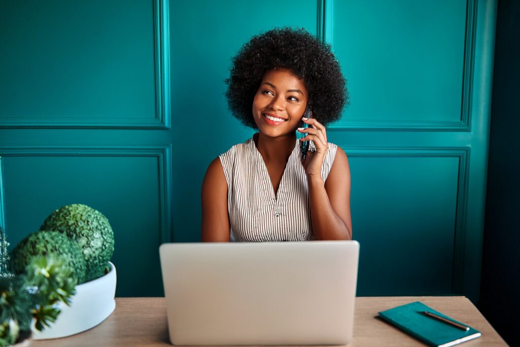 Smiling woman talking on mobile phone while sitting at a laptop in a room with teal coloured walls 